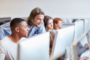 Students looking at desktop screens. A teachers stands over them. 