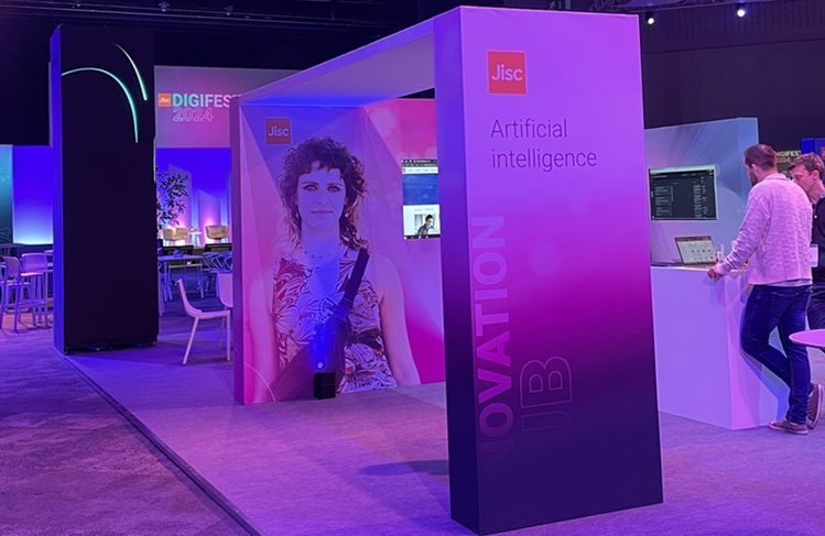 The image shows the Artificial Intelligence stand at Digifest 2024. The stand is inside a conference centre and the light is low, with a pink hue. The stand has text which reads 'Artificial Intelligence' and 'Jisc'. There are two people looking at a laptop on the stand.