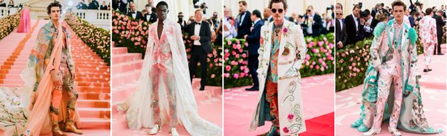 A set of AI generated images showing four male models on a pink carpet, each wearing elaborate, ornate outfits:First: A multicolored suit with an embroidered cape and gold boots. Second: A pink lace suit with a long, white embroidered cape. Third: A white coat with floral embroidery over a colorful patterned outfit. Fourth: A pink suit with teal accents and a matching embroidered cape. Each look is highly detailed, combining traditional tailoring with extravagant, decorative elements.