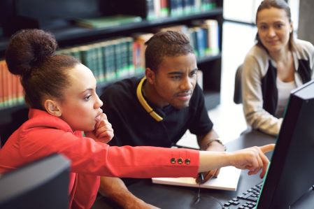 Three students work together at a single computer, the student in the foreground points at the screen and looks thoughtful.