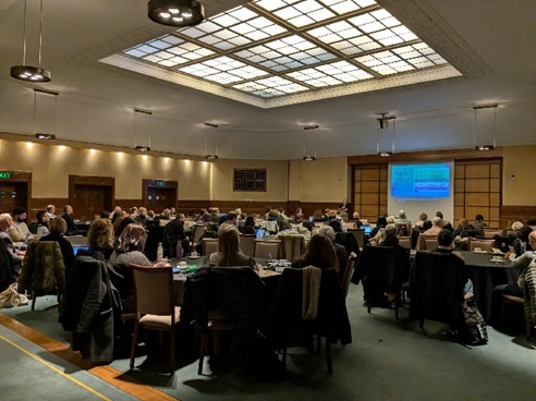 A large conference room filled with attendees seated at round tables, listening to a presentation. The room has warm lighting, with a high ceiling featuring a grid of illuminated panels. A projector screen at the front displays a slide with blue text. Many attendees have laptops open, and some are taking notes. Coats are draped over chairs, indicating a formal yet comfortable setting.