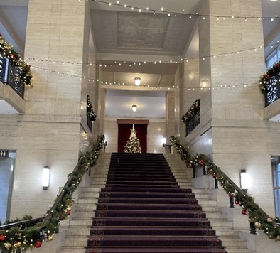 A grand staircase in a well-lit, elegant building, decorated for the holiday season. The staircase is adorned with garlands featuring red and gold ornaments, and string lights are draped across the upper area. A large, decorated Christmas tree stands at the top of the stairs against a red backdrop. The cream-colored walls and high ceiling with ornate detailing enhance the festive and formal atmosphere.