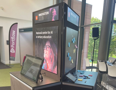 A photograph of Jisc's AI stand for the ALT-C 2023 conference. On one side of the stand is a monitor with the AI demo website. On the other side is a grid with colourful post it notes stuck to it.