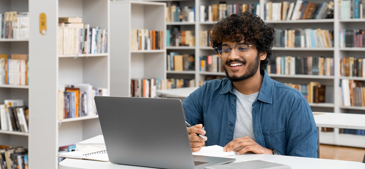 A cheerful man taking notes beside a laptop in a library.