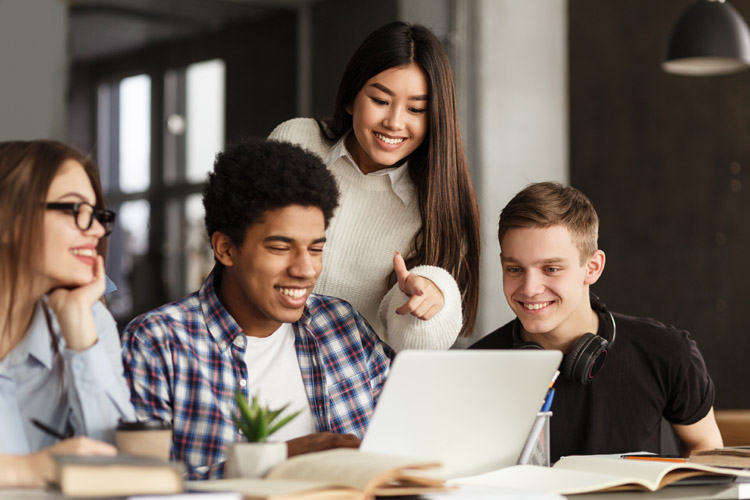Four college students using laptop in library, studying together for school assignment.