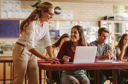 A teacher assisting a smiling student with her laptop in a classroom.