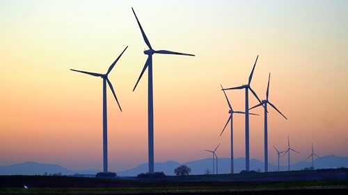 This image shows several large wind turbines standing in an open landscape at sunset. The turbines' blades are rotating gently, with a pastel-colored sky of orange, pink, and blue blending into the horizon. In the background, there are distant hills and some sparse trees, creating a peaceful and serene atmosphere.