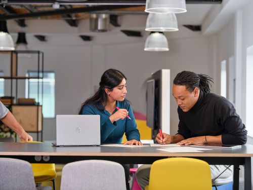Two students study together in an office, one has an open laptop and the other works with pen and paper.