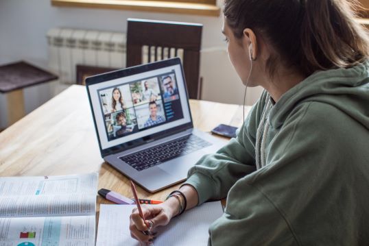 A young woman sits at a wooden table, wearing a green hoodie and earphones, attending a virtual class on her laptop. The laptop screen shows a video call with five participants in a grid format, each in their own environment.