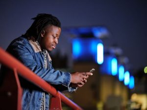 A man focusing on his mobile phone stands at a red railing outside at night.