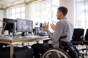 A man in a wheelchair participates in an online meeting, he has two screens showing four attendees on each screen.