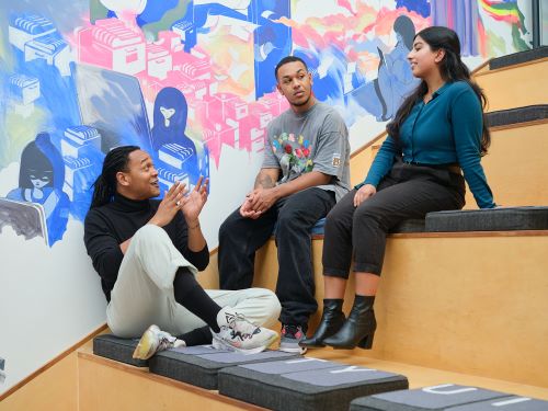 Three students sit on tiered wooden seating, engaged in lively conversation.