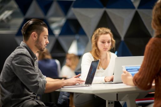 Three individuals are seated at a round table in a modern, collaborative workspace, focused on their laptops.