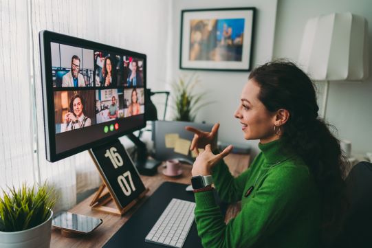 A woman in a green sweater is seated at a desk, engaged in a virtual meeting displayed on a large monitor.