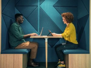 Two people - a man and a woman - sit facing each other in a contemporary study area, one is using a laptop and the other a tablet to work on.