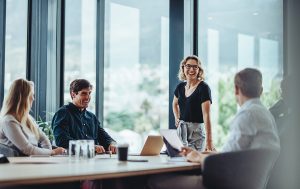 A group of colleagues in a bright meeting room, with one person standing and speaking while others sit around a table.